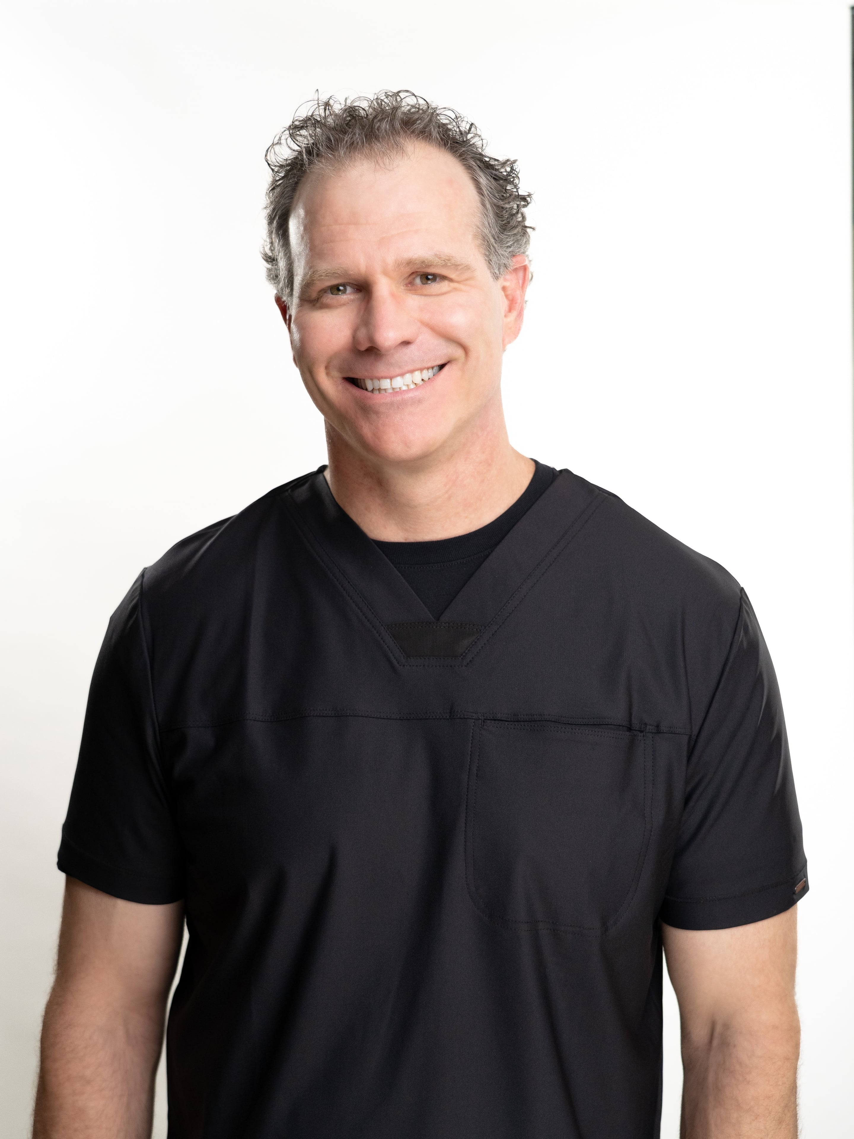 Bryce Wolcott wearing black medical scrubs over a black undershirt, photographed against a plain white studio background.