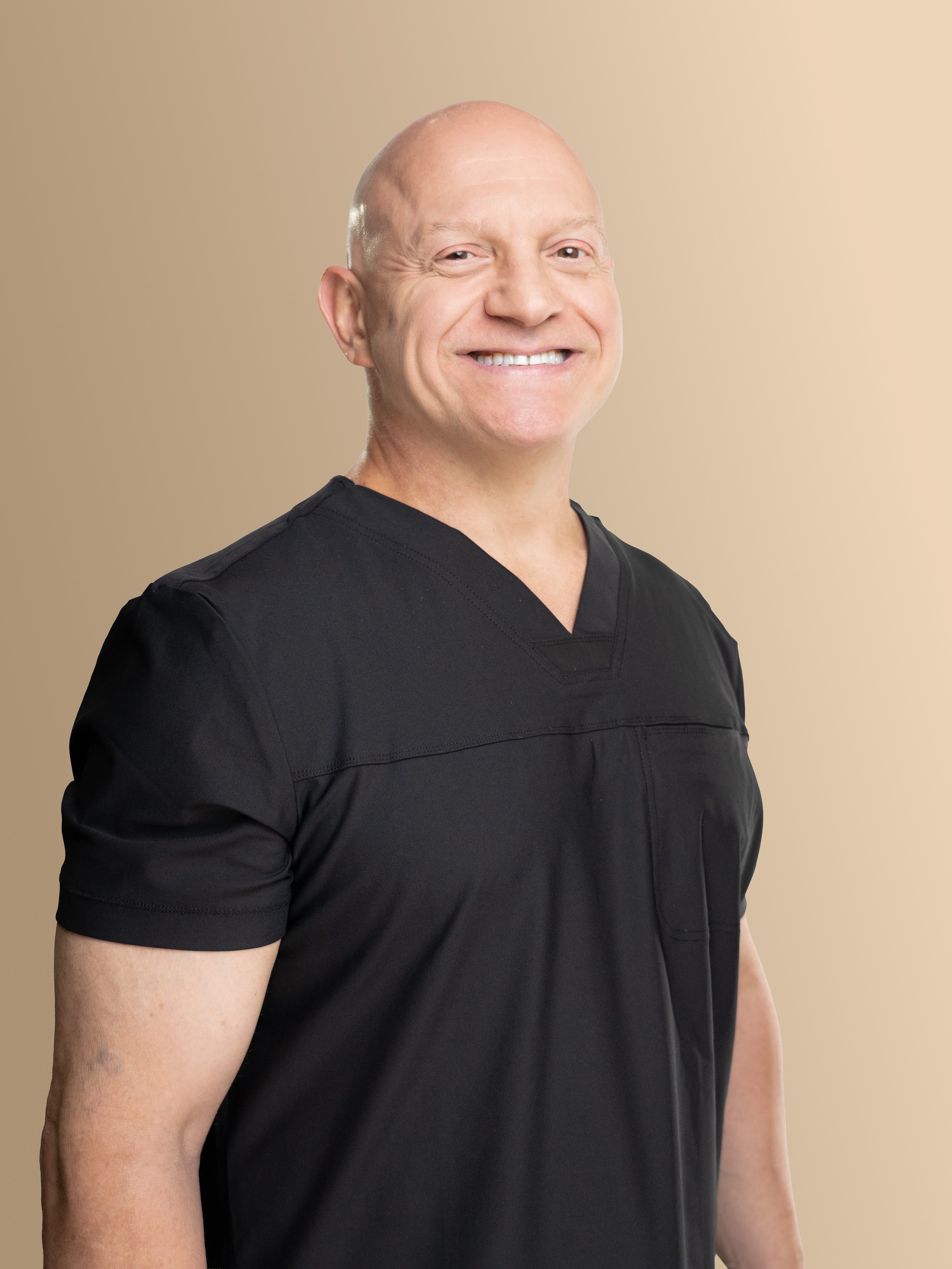 Dr. Thomas Incledon wearing a black V-neck scrub top, photographed against a plain beige studio background.