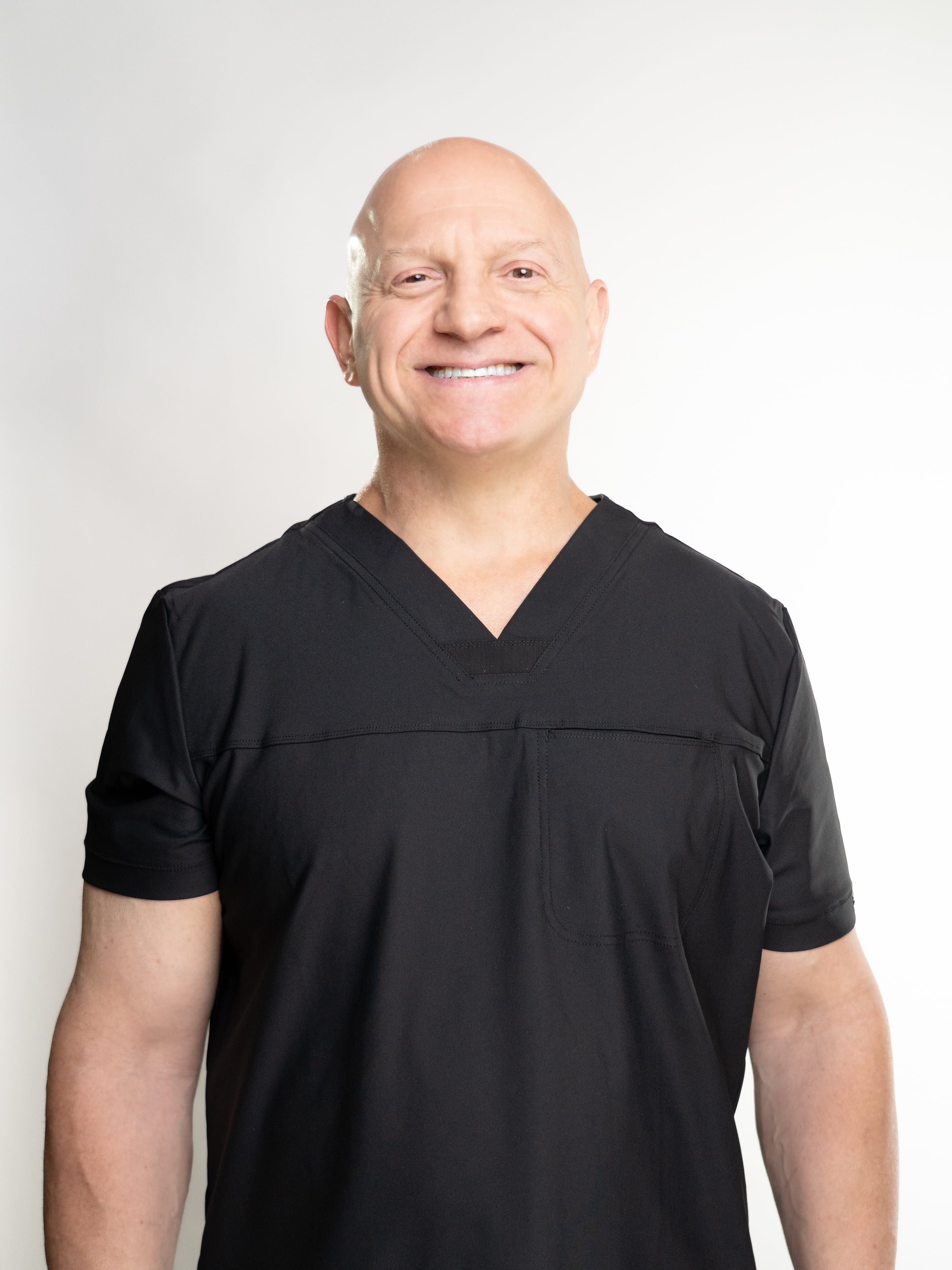 Dr. Thomas Incledon wearing a black V-neck scrub top, photographed against a plain light gray studio background.
