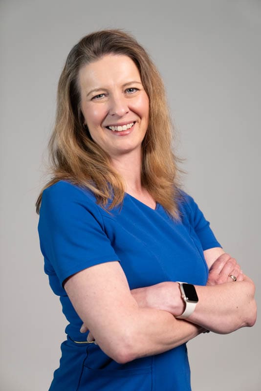Dr. Kristy Anderson wearing blue crubs with arms crossed on a gray background