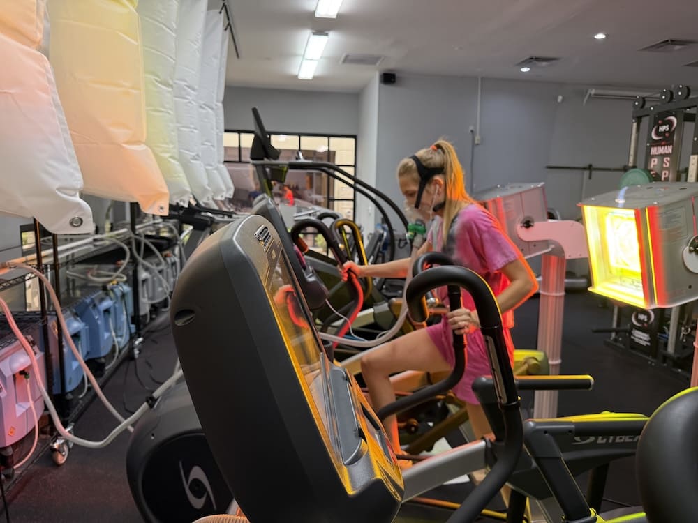 Woman using an EWOT machine in a gym setting with colorful equipment and a brick wall.