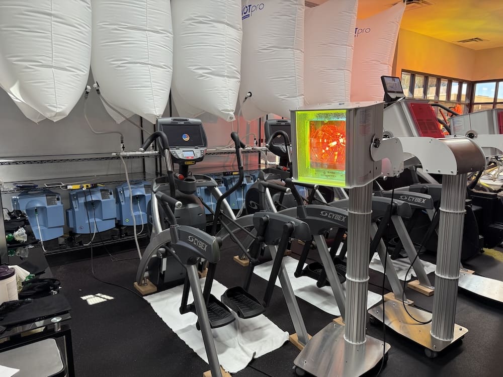 A fitness training setup featuring multiple Cybex elliptical exercise machines in a gym-like room with large white inflatable bags suspended overhead and blue storage containers along the left wall.
