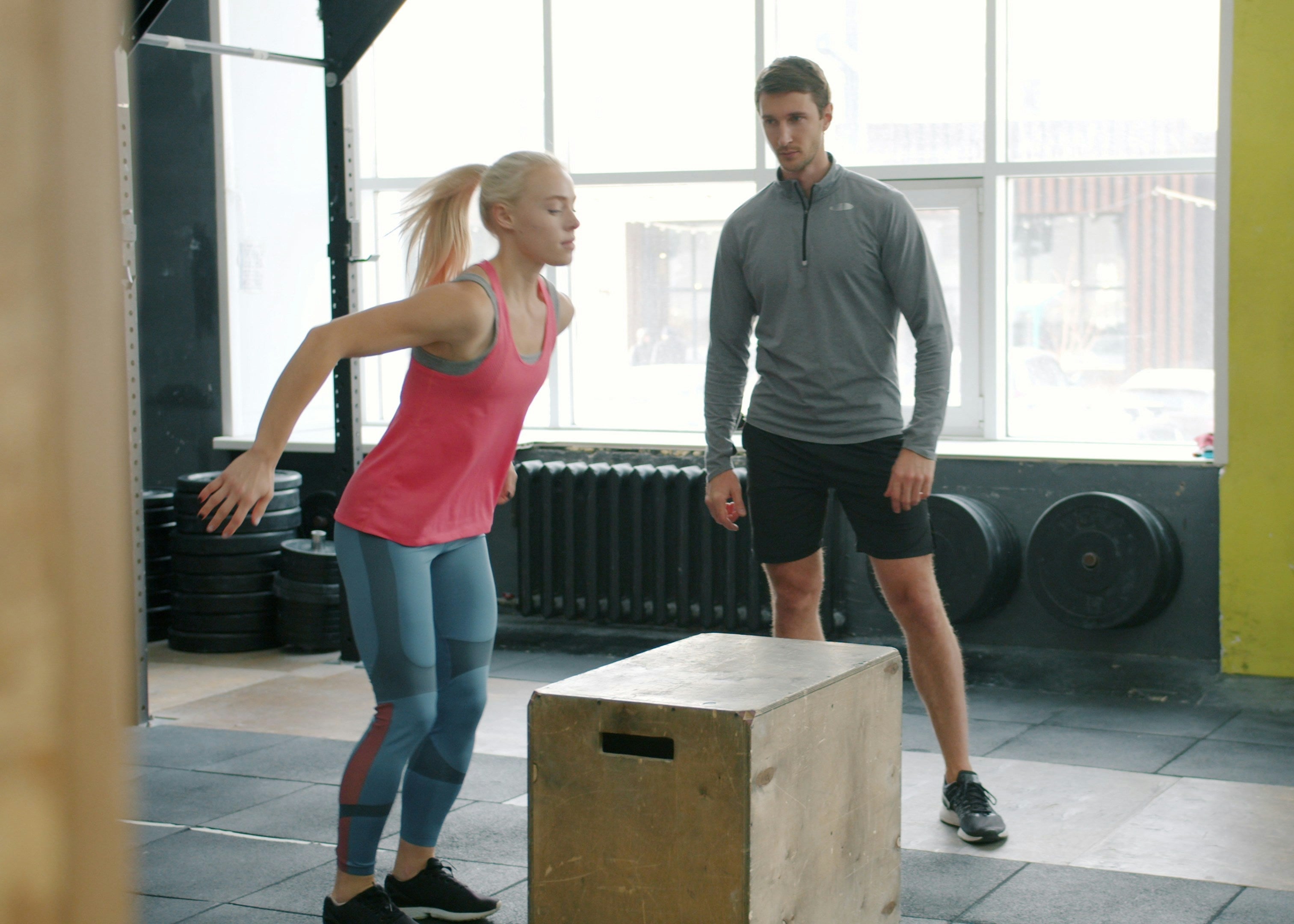 A man and a woman in a gym setting, with the woman in a pink tank top and blue leggings preparing to jump onto a wooden plyo box while the man in a gray long-sleeve shirt and black shorts watches.